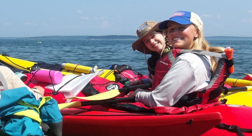 Two people wearing life jackets sit inside colorful kayaks and smile for the photo. 